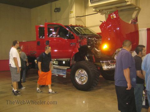 1990 Ford Bronco - large red medium-duty pickup truck with its hood raised is displayed on a polished floor while a crowd of spectators looks it over indoors.