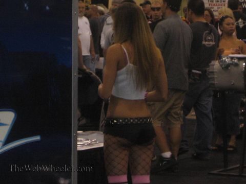 1990 Ford Bronco - Rear view of a woman in a white tank top, dark shorts and pink-cuffed fishnets standing in a crowded indoor motorsports expo aisle.