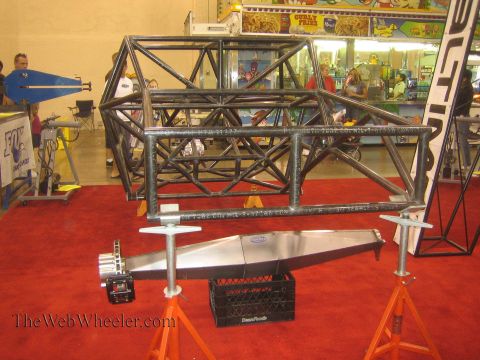 1990 Ford Bronco - welded steel space-frame tube chassis section is displayed on jack stands above polished aluminum parts on red carpet at an indoor motorspo…