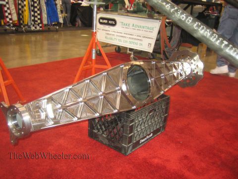 1990 Ford Bronco - polished aluminum truss-style chassis tube with triangulated lightening panels rests on a black milk crate over red carpet at an indoor exp…