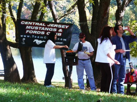 2001 Mercury Grand Marquis - A group of people enjoy a picnic and barbecue under trees, with a club banner in the background.