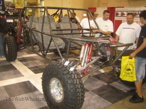 1990 Ford Bronco - unpainted tube-frame off-road buggy chassis with multiple seats, coilover shocks and a beadlock-wheeled mud tire is shown to onlookers at a…