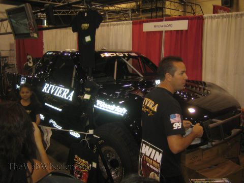 1990 Ford Bronco - glossy black race truck lettered RIVIERA fills a booth while a man in a Riviera Racing shirt stands in the foreground at an indoor expo.