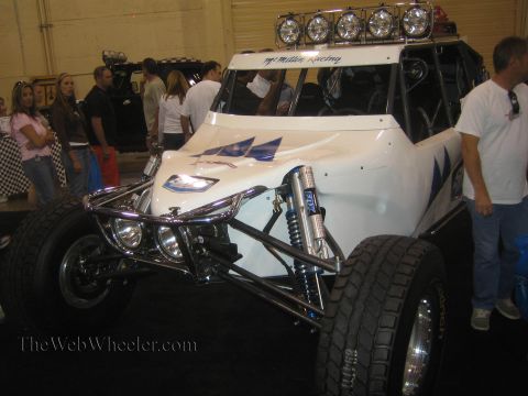 1990 Ford Bronco - white single-seat off-road race buggy with a five-light roof bar, exposed front A-arms and coilover shocks sits among a crowd at an indoor…