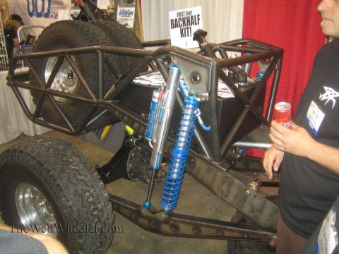 1990 Ford Bronco - rear half of an unpainted tube-frame buggy with blue coilover shocks and mud tires sits beside a BACKHALF KIT sign as a man holds a soda ca…