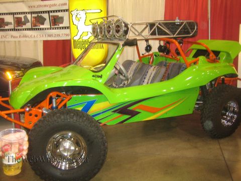 1990 Ford Bronco - bright green two-seat off-road buggy with an orange tube frame, roof-mounted spare tires and graphic decals sits on display at an indoor ex…