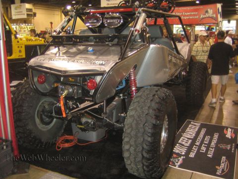 1990 Ford Bronco - bare silver tube-frame rock buggy with a front winch, coilover shocks and oversized mud tires sits beside a display sign at an indoor expo.