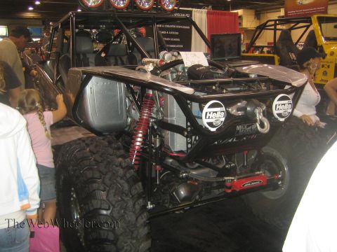 1990 Ford Bronco - rear of a black tube-frame rock buggy fitted with two round Hella lights, a coilover shock and oversized tire draws a crowd at an indoor ex…