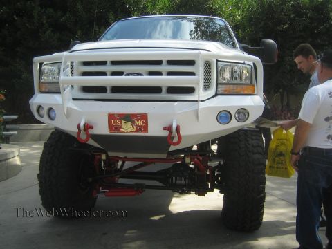 1990 Ford Bronco - white lifted Ford Super Duty pickup shows its custom front bumper, grab hooks and solid front axle as a man stands beside it outdoors.