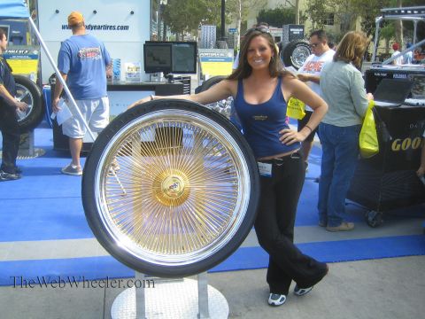 1990 Ford Bronco - smiling woman in a blue tank top leans on a tall gold wire-spoke wheel mounted on a stand at an outdoor Goodyear tire display.