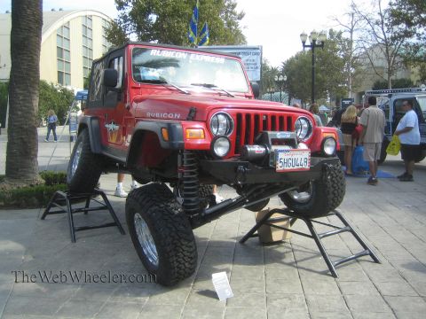 1990 Ford Bronco - red Jeep Wrangler Rubicon with a lift kit and mud tires sits raised on jack stands on a paved plaza at an outdoor event.