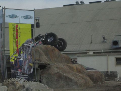1990 Ford Bronco - tube-frame rock buggy climbs nearly vertical up an artificial rock pile next to a UROC event banner outside a metal building.