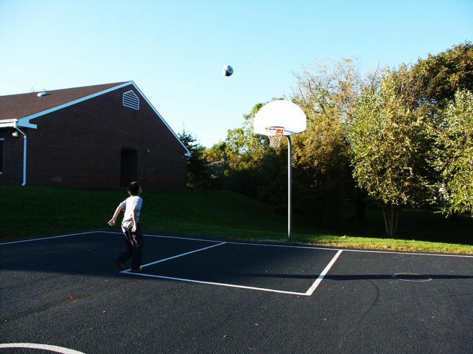 Basketball hoop and court with a boy playing, a brick building, and trees under a clear blue sky