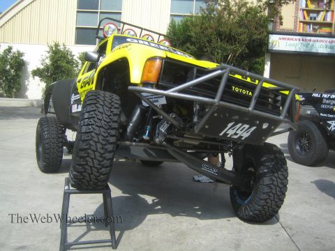 1990 Ford Bronco - yellow and black tube-bed Toyota prerunner race truck numbered 1494 sits raised on a jack stand on pavement outside a building.