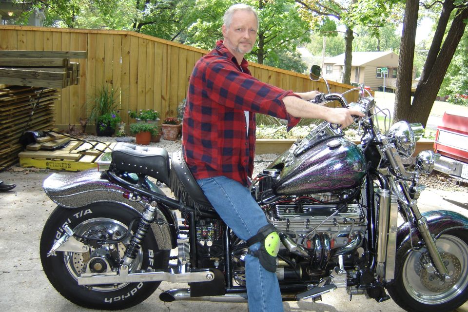 Man on custom chrome motorcycle with purple flame tank and fat BFGoodrich rear tire on a driveway.