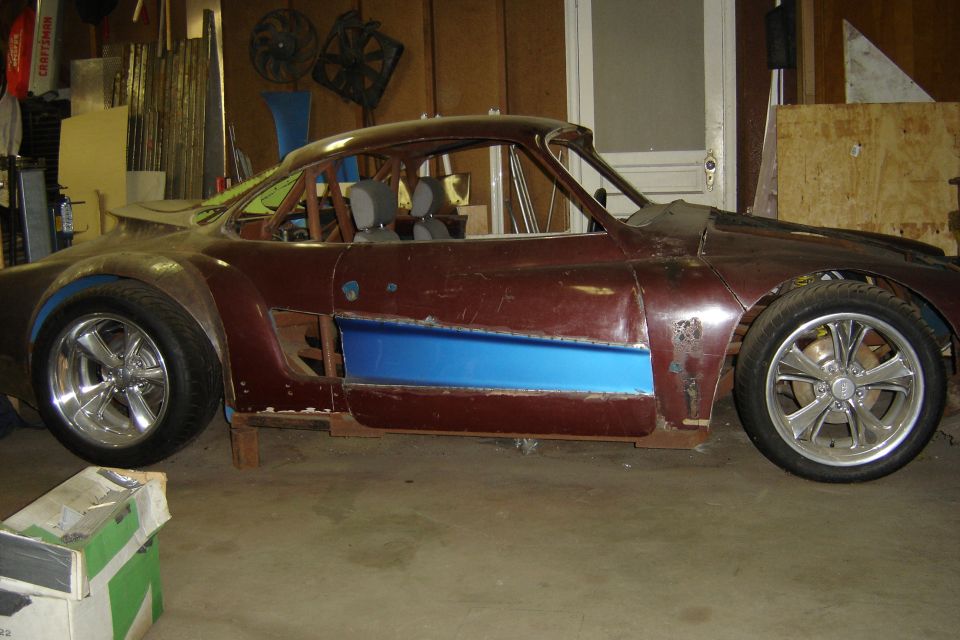 Side view of maroon project coupe with blue door panel, polished wheels, and roll cage, up on blocks in a garage