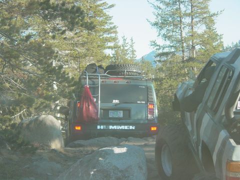 1986 Ford Bronco - A Hummer H2 is seen from the rear on a rocky trail, equipped with a roof rack and spare tire.