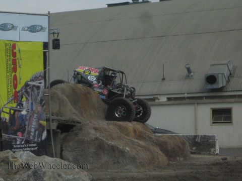 1990 Ford Bronco - black tube-chassis rock buggy marked 123 works up a rocky obstacle in front of a yellow UROC banner and metal building.