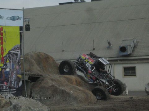 1990 Ford Bronco - black rock-crawling buggy noses down a rocky obstacle into a dirt arena past a UROC banner.