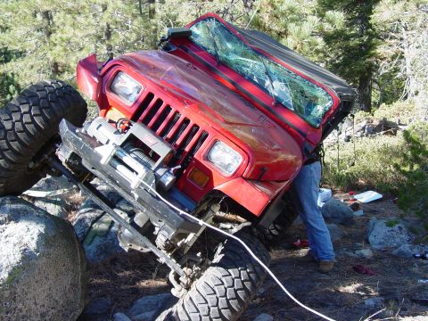 1986 Ford Bronco - A red Jeep Wrangler is stuck on rocks, its windshield shattered, with a person attending to it.