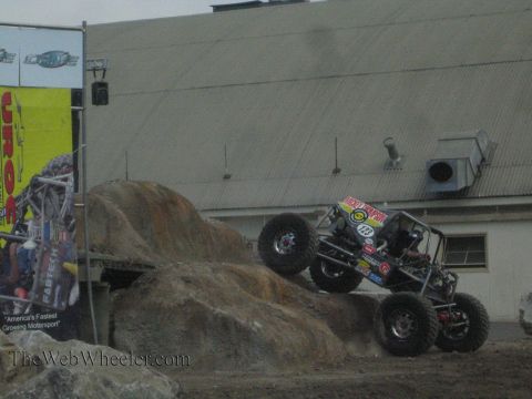 1990 Ford Bronco - black tube-frame rock buggy descends a boulder onto dirt at a UROC rock-crawling competition.