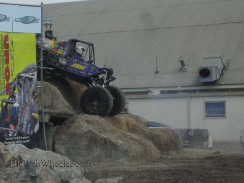 1990 Ford Bronco - purple rock-crawling buggy with AEM livery crawls up a steep rock face past a UROC banner.
