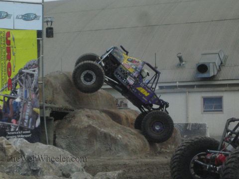 1990 Ford Bronco - purple AEM-liveried rock buggy flexes over boulders as a second tube buggy waits at right.