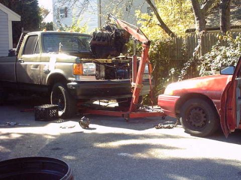 1979 Ford Bronco - A green pickup truck is parked next to a red car, with its engine being lifted by a crane.