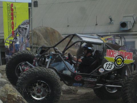 1990 Ford Bronco - Side view of a black Mickey Thompson rock buggy number 123 with its driver seated, parked by boulders at a UROC event.