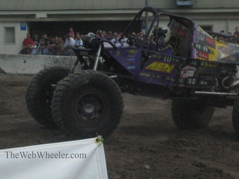1990 Ford Bronco - purple AEM-liveried tube rock buggy with a helmeted driver sits in a dirt arena before a crowd of spectators.