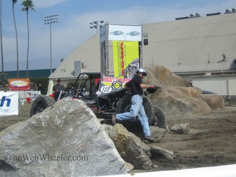 1990 Ford Bronco - crew member pushes the rear of a black rock buggy number 123 over boulders in an outdoor dirt course with palm trees.