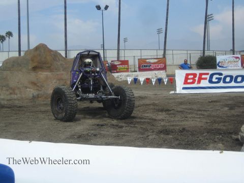 1990 Ford Bronco - blue tube-chassis rock buggy with a helmeted driver sits on a dirt course near a BFGoodrich banner and spectators.