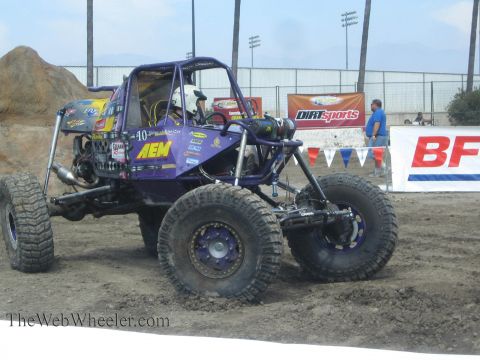 1990 Ford Bronco - purple AEM-liveried tube rock buggy with beadlock wheels sits on dirt beside DirtSports and BFGoodrich banners.