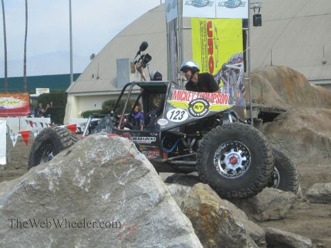 1990 Ford Bronco - black rock buggy number 123 with Mickey Thompson branding crawls over big boulders while a videographer films nearby.