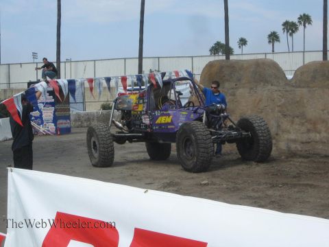 1990 Ford Bronco - purple AEM-liveried tube rock buggy sits on a dirt course with crew members and palm trees in the background.