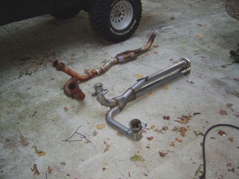 1986 Ford Bronco - Two exhaust assemblies laid on a concrete garage floor: a rusty stock Y-pipe with catalytic converter on the left and a shiny stainless-ste…