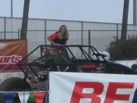 1990 Ford Bronco - smiling blonde woman in an orange and black shirt poses behind a Maxxis-sponsored off-road tube buggy near event banners.