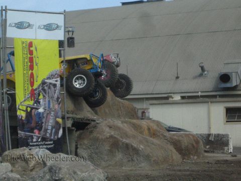 1990 Ford Bronco - yellow and blue tube-chassis rock buggy climbs a steep boulder obstacle beside a yellow UROC off-road challenge banner.
