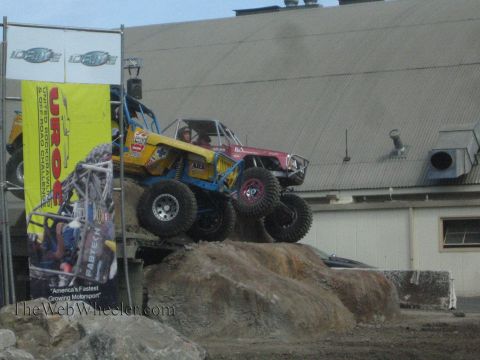 1990 Ford Bronco - yellow-blue buggy and a red buggy navigate a large boulder obstacle in front of a yellow UROC banner against a metal building.