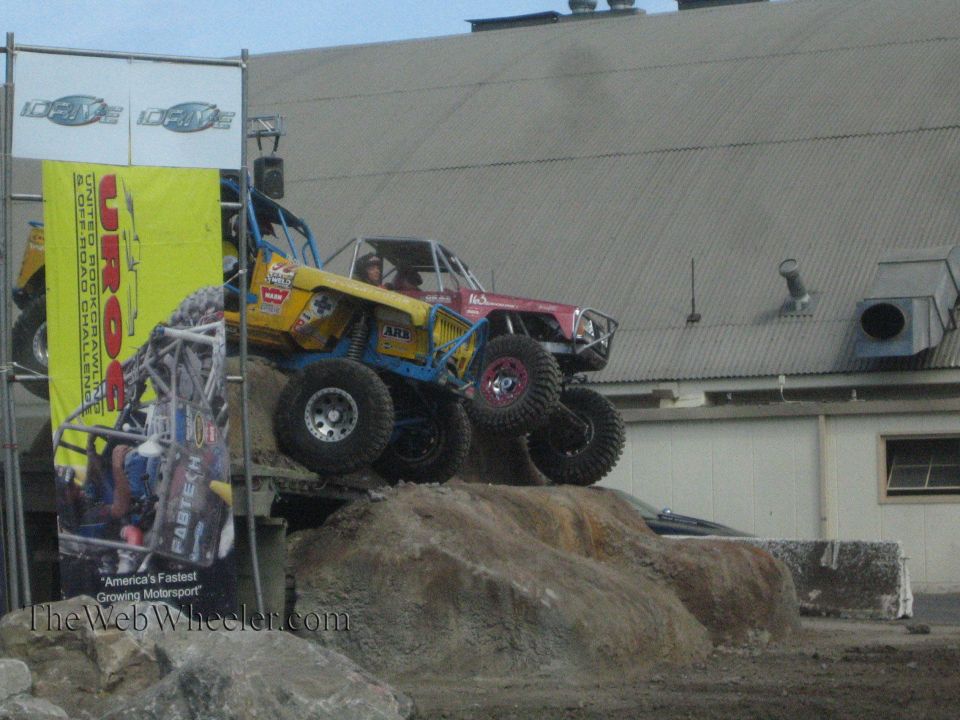 Yellow buggy and red buggy on a rock obstacle near a UROC banner at an outdoor event