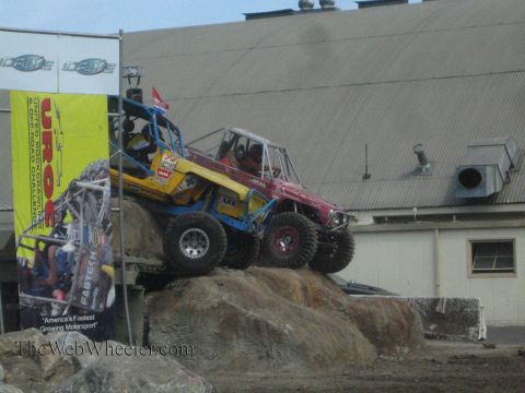 1990 Ford Bronco - yellow and blue tube-chassis rock buggy crawls over a large rock obstacle next to a UROC banner with a red buggy behind.