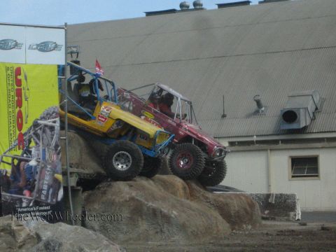 1990 Ford Bronco - yellow-blue rock buggy and a red rock buggy climb a man-made boulder pile near a UROC banner and metal warehouse.