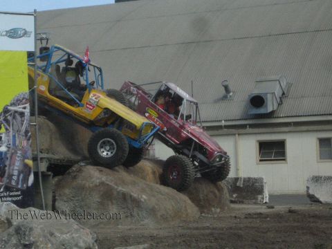 1990 Ford Bronco - yellow-blue tube buggy and a red tube buggy descend a steep boulder obstacle in front of a metal warehouse building.