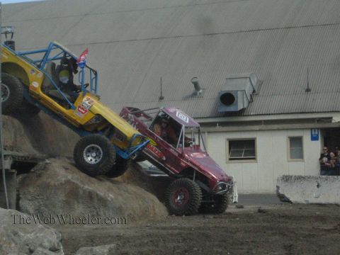 1990 Ford Bronco - yellow-blue rock buggy and a maroon rock buggy descend a boulder obstacle as spectators watch beside a warehouse building.
