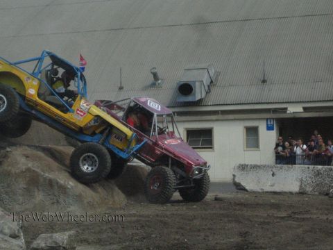 1990 Ford Bronco - yellow-blue tube buggy and a maroon tube buggy work a boulder obstacle while a crowd of spectators watches behind a barrier.