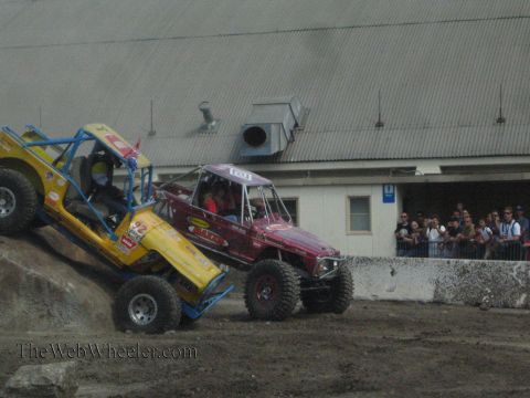 1990 Ford Bronco - yellow-blue rock buggy and a maroon rock buggy work a dirt and rock obstacle as spectators line a fence near a warehouse.