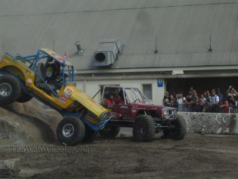 1990 Ford Bronco - yellow-blue rock buggy climbs a rock ledge while a maroon buggy sits alongside and a crowd of spectators watches.