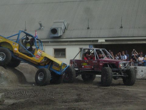 1990 Ford Bronco - yellow-blue rock buggy and a maroon rock buggy sit side by side on a dirt course as spectators watch from behind a fence.