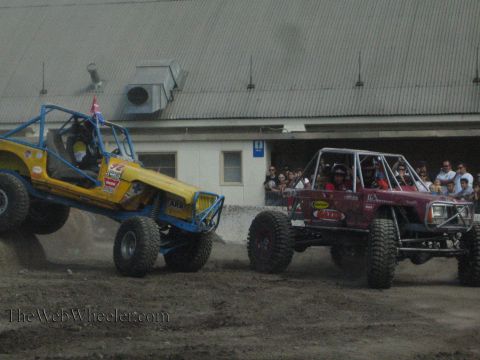 1990 Ford Bronco - yellow-blue rock buggy and a maroon rock buggy drive across a dusty dirt course as spectators line a fence near a warehouse.