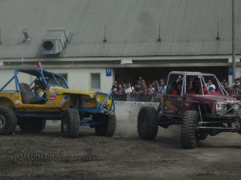 1990 Ford Bronco - yellow-blue rock buggy and a maroon rock buggy maneuver on a dusty dirt course as a crowd of spectators watches behind a barrier.